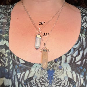 May include: Two silver-toned necklaces with crystal point pendants. The top necklace features a light-colored crystal and is labeled 20 inches, while the bottom necklace features a tan-colored crystal and is labeled 22 inches in length.