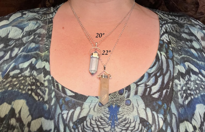 May include: Two silver-toned necklaces with crystal point pendants. The top necklace features a light-colored crystal and is labeled 20 inches, while the bottom necklace features a tan-colored crystal and is labeled 22 inches in length.