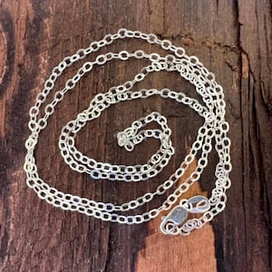 May include: A silver chain necklace featuring small, uniform oval-shaped links connected in a continuous loop. The chain includes a polished lobster claw clasp mechanism at one end, designed for secure fastening, with a bright, reflective metallic finish.