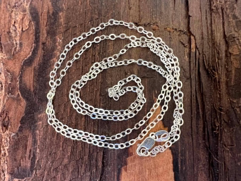May include: A silver chain necklace featuring small, uniform oval-shaped links connected in a continuous loop. The chain includes a polished lobster claw clasp mechanism at one end, designed for secure fastening, with a bright, reflective metallic finish.
