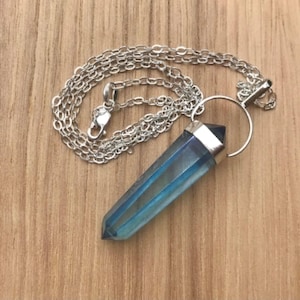 May include: A pendant necklace featuring a faceted, elongated blue crystal point suspended from a silver-toned metal bail and a delicate silver-toned chain. The crystal displays a translucent, iridescent finish with deep blue hues and a pointed tip.
