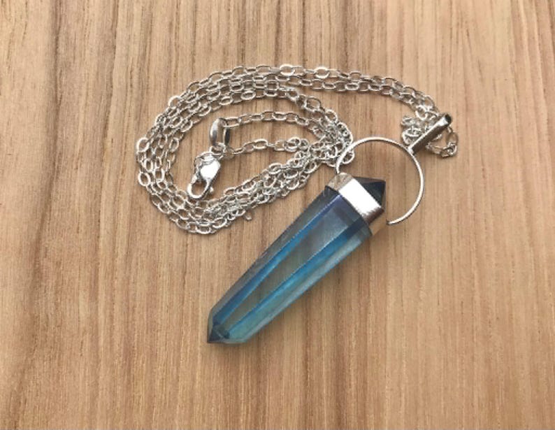 May include: A pendant necklace featuring a faceted, elongated blue crystal point suspended from a silver-toned metal bail and a delicate silver-toned chain. The crystal displays a translucent, iridescent finish with deep blue hues and a pointed tip.