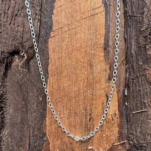 May include: A delicate silver-toned metal chain necklace featuring a series of small, uniform circular links. The chain is presented in a simple, continuous loop design, showcasing a bright, reflective metallic finish against a textured background.