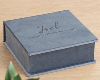 Op de afbeelding: Een vierkante bewaardoos van leigrijs fluweel met een scharnierend deksel. De bovenkant van de doos is gegraveerd met de naam "Joel" in een cursief lettertype, gevolgd door de tekst "June 2010 - November 21, 2020" in een strak schreefloos lettertype.