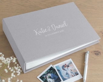 May include: A gray fabric-covered wedding photo album with the names "Katie & Daniel" and the date "30 November 2020" printed on the cover. Two polaroid photos and a pen are next to the album. The album is open, revealing white pages.