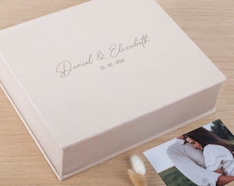 May include: A square, light beige keepsake box with a hinged lid. The top of the box features the printed text "Daniel & Elizabeth" in a cursive script font, followed by the date "21.10.2021" in a simple, clean sans-serif typeface.
