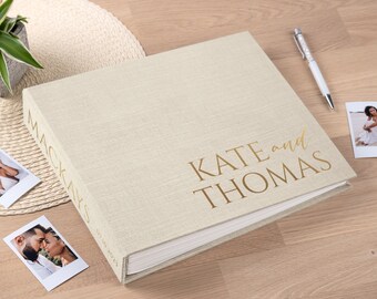 May include: A beige linen-covered photo album with gold lettering that reads "KATE and THOMAS." The spine reads "MACKAYS" and the date "07.10.2023." Two photos and a pen are next to the album. A small plant in a pot is in the background.