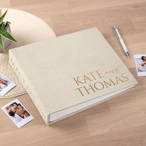 May include: A beige linen-covered photo album with gold lettering that reads "KATE and THOMAS." The spine reads "MACKAYS" and the date "07.10.2023." Two photos and a pen are next to the album. A small plant in a pot is in the background.