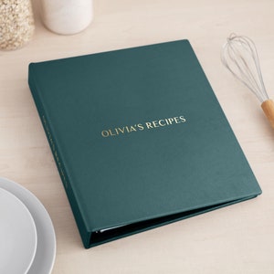 May include: A dark teal recipe binder with a textured cover featuring the gold embossed text OLIVIA'S RECIPES on the front and the spine. The binder is closed and rectangular, showcasing a clean, solid-colored surface with metallic lettering.