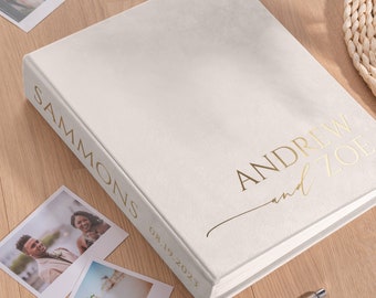 May include: A rectangular photo album with a cream-colored cover featuring gold foil text that reads "SAMMONS" on the spine and "ANDREW and ZOE" with the date "08.19.2023" on the front. The album is surrounded by several small square photographs.