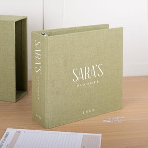 Puede incluir: Una carpeta de tres anillas de color verde salvia con una cubierta de tela texturizada. El frente y el lomo presentan el texto blanco SARA'S PLANNER y el año 2025. Una funda a juego de color verde salvia se sitúa a la izquierda.