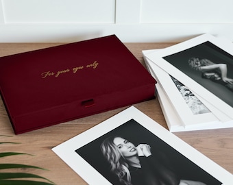 May include: A rectangular burgundy storage box with a small ribbon pull tab and the gold embossed text For your eyes only on the lid, accompanied by several black and white photographic prints with white borders displayed on a wooden surface.