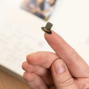Pode incluir: Um pequeno autocolante de um chap&eacute;u alto preto com uma faixa amarela. O autocolante &eacute; segurado entre dois dedos, com um fundo desfocado de um livro e uma foto.