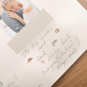 Pode incluir: Uma p&aacute;gina de scrapbook aberta com uma fotografia de um casal. A p&aacute;gina apresenta texto manuscrito e ilustra&ccedil;&otilde;es de um chap&eacute;u, an&eacute;is e um cupido. O texto diz "To the moon and back! Lucy Congratulations :) sleep well."