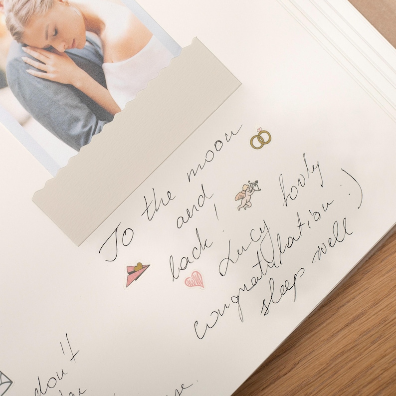 Pode incluir: Uma p&aacute;gina de scrapbook aberta com uma fotografia de um casal. A p&aacute;gina apresenta texto manuscrito e ilustra&ccedil;&otilde;es de um chap&eacute;u, an&eacute;is e um cupido. O texto diz "To the moon and back! Lucy Congratulations :) sleep well."