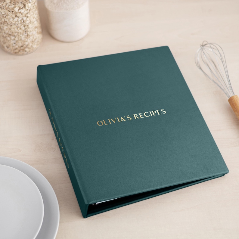 May include: A dark teal recipe binder with a textured cover featuring the gold embossed text OLIVIA'S RECIPES on the front and the spine. The binder is closed and rectangular, showcasing a clean, solid-colored surface with metallic lettering.