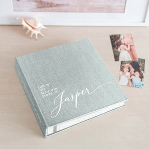 May include: A light blue linen-covered photo album with the text "THIS IS THE BEAUTIFUL STORY OF Jasper" in white script. Two photos and a seashell are on the light wood surface.