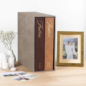 Peut inclure: Un coffret de rangement taupe contenant deux albums photo verticaux aux couvertures marron foncé et beige. Chaque dos porte le texte doré Fosters et les chiffres romains I et II. Un cadre photo doré et des bougies blanches sont présents.