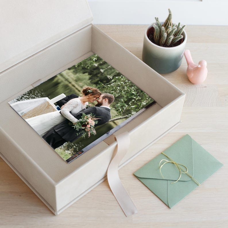 May include: A large, light-coloured, rectangular keepsake box containing a photograph of a couple embracing outdoors. The box features a ribbon pull tab. Nearby sits a small green succulent in a gradient pot, a pink bird figurine, and a green envelope.