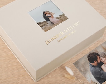 Op de afbeelding: Een vierkante, crèmekleurige fotoalbumdoos met een centrale vierkante foto van een koppel dat elkaar buiten omhelst. De tekst JENNIFER & HENRY en JANUARY 18 2025 is in goudfolie op de omslag gedrukt. Een losse, bijpassende foto ligt ernaast.