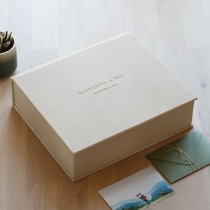 May include: A rectangular off-white keepsake box with a hinged lid featuring the embossed text MACKENZIE & BEN and OCTOBER 11, 2023. The box is positioned near a small green envelope tied with gold string and a photograph of two people in a landscape.