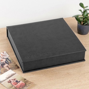 Puede incluir: Una caja cuadrada negra con acabado texturizado y el texto BODE ICKE en relieve en la tapa. La caja está colocada sobre una superficie de madera junto a dos fotografías rectangulares y una pequeña planta verde en una maceta gris oscuro.