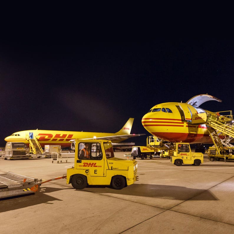 May include: Two yellow DHL cargo planes are parked on the tarmac at night. The plane on the right has its cargo door open and a yellow baggage tug is pulling a luggage cart towards the open door.