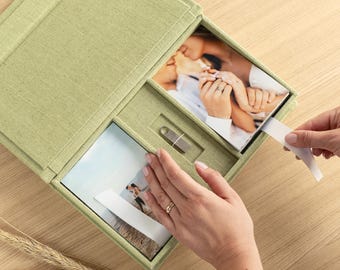 May include: A light green fabric-covered presentation box featuring a hinged lid, designed to hold photographic prints and a small silver USB flash drive. The interior contains two recessed compartments for prints and a central slot for the drive.
