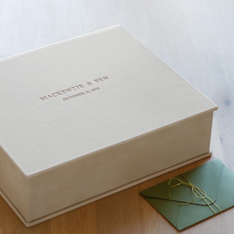 May include: A rectangular, off-white keepsake box with a smooth, matte finish. The lid features the embossed text MACKENZIE & BEN and OCTOBER 31, 2023. Beside the box lies a small, olive green envelope tied with a thin, metallic gold string.