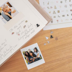Pode incluir: Um &aacute;lbum de casamento aberto com uma foto de um casal e um polaroid de amigos. H&aacute; texto manuscrito e adesivos decorativos. Mais adesivos e uma superf&iacute;cie de madeira est&atilde;o espalhados.