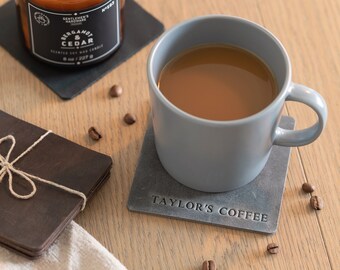 May include: A grey ceramic coffee mug with a brown coffee drink inside, sitting on a grey square coaster with the text "TAYLOR'S COFFEE" printed on it. The mug is on a wooden table with coffee beans scattered around it.
