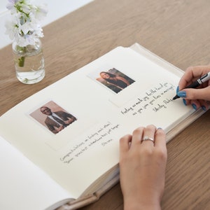 Puede incluir: Un &aacute;lbum de fotos abierto con dos fotos enmarcadas y mensajes manuscritos. Una persona escribe con un bol&iacute;grafo negro en la p&aacute;gina derecha. Un peque&ntilde;o jarr&oacute;n con flores blancas se encuentra en el fondo sobre una mesa de madera.