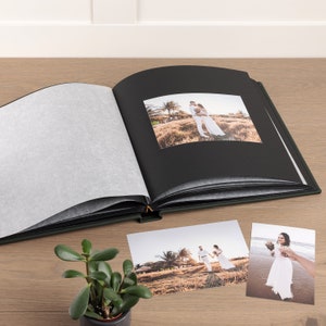 May include: An open, dark green photo album with black pages and a gold clasp. The album contains a wedding photo, with two additional photos of a couple nearby. A small potted plant sits in the foreground on a wooden surface.