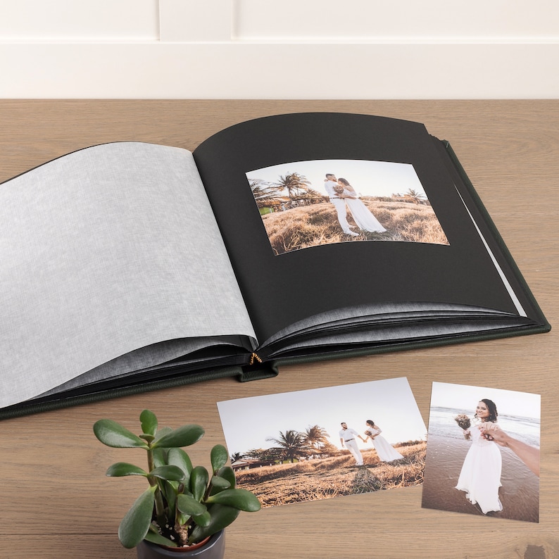 May include: An open, dark green photo album with black pages and a gold clasp. The album contains a wedding photo, with two additional photos of a couple nearby. A small potted plant sits in the foreground on a wooden surface.