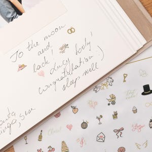 Pode incluir: Um close-up de um cart&atilde;o branco com texto manuscrito e pequenas ilustra&ccedil;&otilde;es. O texto diz: "To the moon and back! Lucy lovely! Congratulation :) sleep well". Os autocolantes decorativos incluem cora&ccedil;&otilde;es, um bolo e uma garrafa de champanhe.