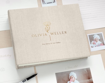 May include: A beige linen baby memory book with gold foil lettering that reads "OLIVIA WELLER" and "the story of our baby." The book is surrounded by baby photos and stationery. A pen is in the foreground.