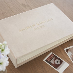 Puede incluir: Un &aacute;lbum de fotos rectangular color crema con los nombres "ANDREW & MEGAN" y la fecha "11.12.2021" en letras doradas. El &aacute;lbum est&aacute; sobre una superficie de madera, con algunas fotos impresas cerca.
