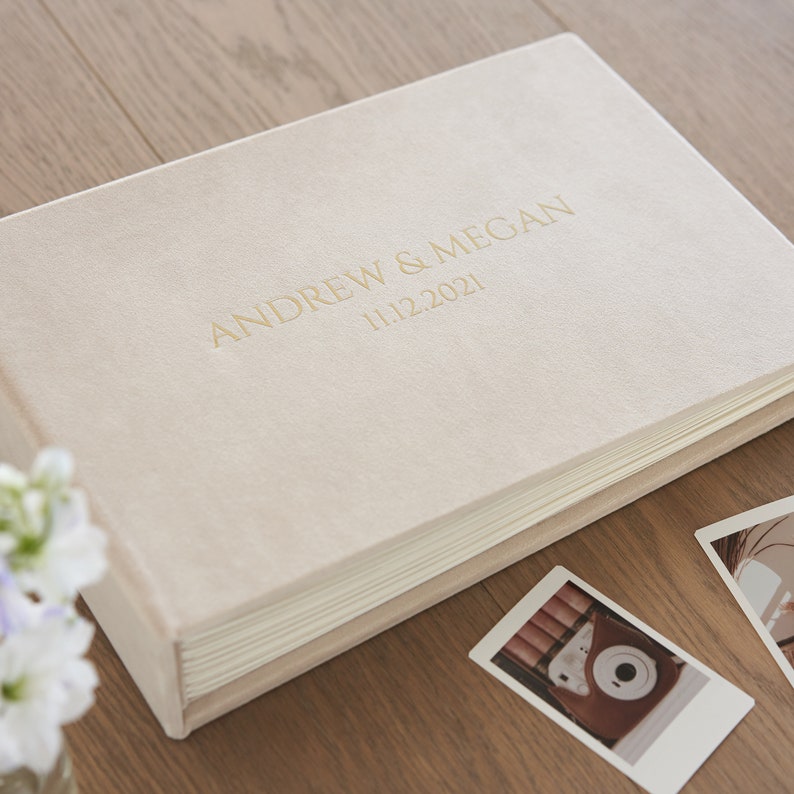 Puede incluir: Un &aacute;lbum de fotos rectangular color crema con los nombres "ANDREW & MEGAN" y la fecha "11.12.2021" en letras doradas. El &aacute;lbum est&aacute; sobre una superficie de madera, con algunas fotos impresas cerca.