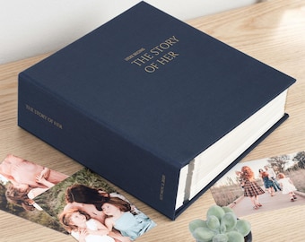 May include: A navy blue photo album with gold foil text on the cover that reads "HERE BEGINS THE STORY OF HER". The spine features the text "THE STORY OF HER" and "EST NOV 2 2020". The album is square with white pages visible.