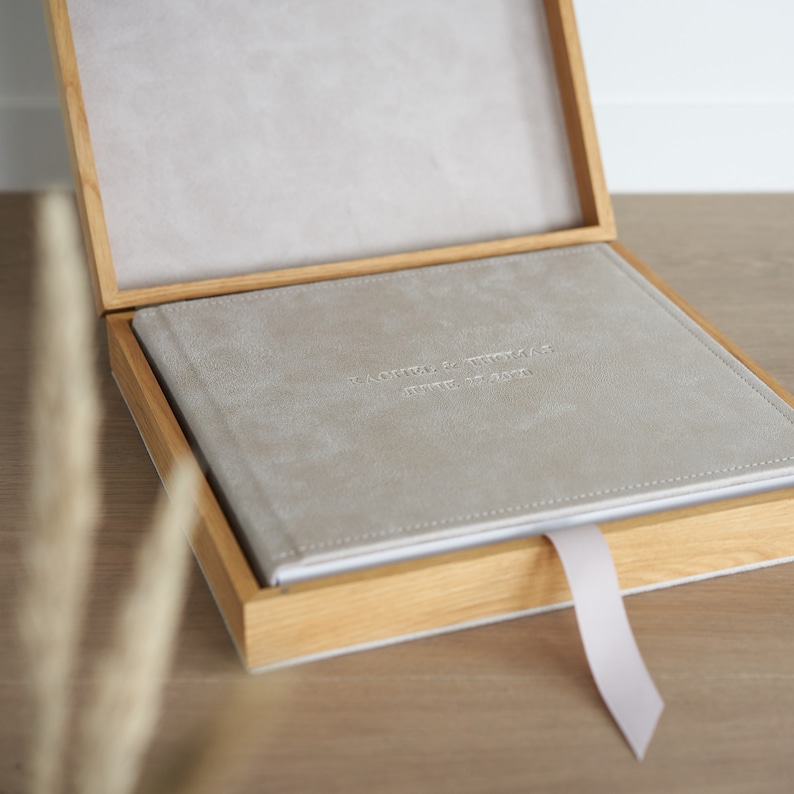 May include: A light gray photo album with embossed text inside a wooden box. The album has a matching light gray interior and a light pink ribbon. The wooden box has a natural wood grain finish. The album's text reads "RACHEL & THOMAS" and "JUNE 27, 2020".