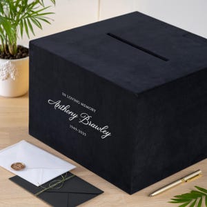 May include: A black velvet box with a slot on top, inscribed with "IN LOVING MEMORY" and the name "Anthony Brawley" along with the years 1949-2025. Envelopes and a gold pen are next to the box.