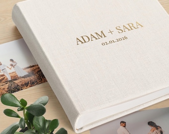 May include: A square photo album with a textured off-white linen cover. The front features gold foil lettering that reads ADAM + SARA and the date 02.01.2026. The album is positioned next to two printed photographs and a small green succulent plant.