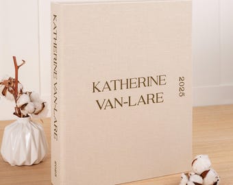 Könnte beinhalten: Ein beiges, mit Leinen bezogenes Buch mit dem Namen "KATHERINE VAN-LARE" und dem Jahr "2025" in dunkelbrauner Schrift. Das Buch steht aufrecht auf einer hellen Holzoberfläche. Links befindet sich eine kleine Vase mit Baumwollstielen.