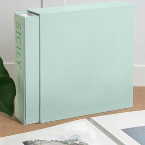 Op de afbeelding: Een licht mintgroene, met stof beklede cassette en boekset. De rug van het boek bevat het woord SICILY in grote groene hoofdletters, met de tekst ANDREA & THOMAS verticaal gedrukt in kleinere groene letters onder de titel.