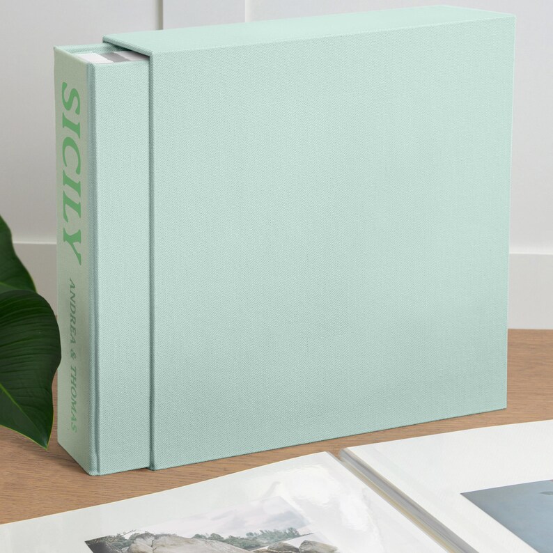 Op de afbeelding: Een licht mintgroene, met stof beklede cassette en boekset. De rug van het boek bevat het woord SICILY in grote groene hoofdletters, met de tekst ANDREA & THOMAS verticaal gedrukt in kleinere groene letters onder de titel.