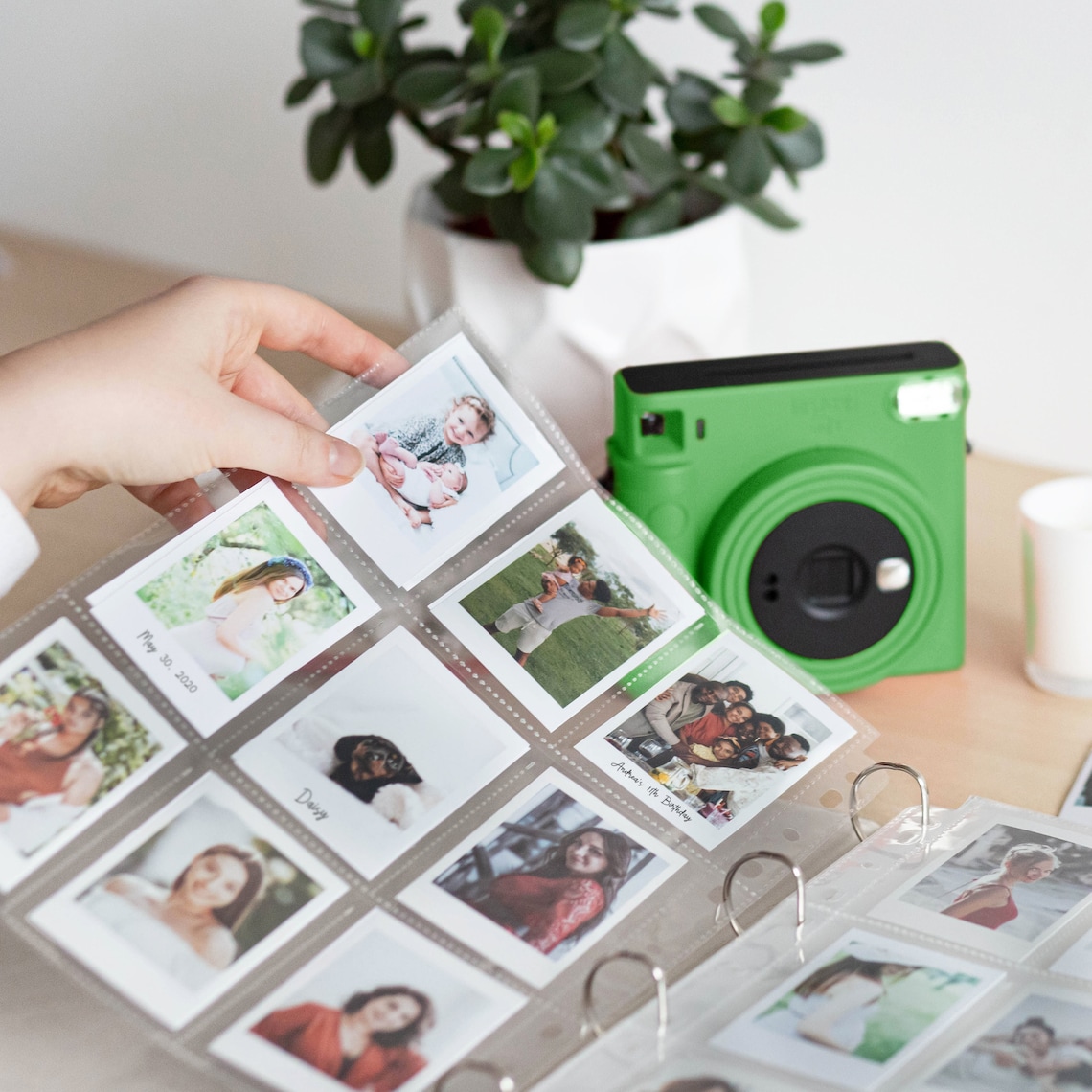 Op de afbeelding: Een groene instant camera met een zwarte lens en een doorzichtig plastic fotoalbum gevuld met instant foto's. Het album heeft een ringband en is gevuld met foto's van mensen en plaatsen.