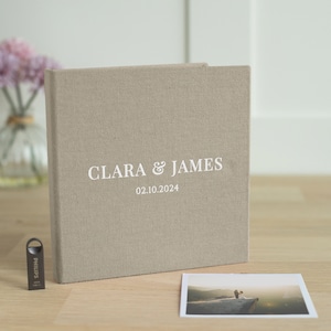 Peut inclure: Un album photo beige en lin avec les noms "CLARA & JAMES" et la date "02.10.2024" imprimés en blanc. Une petite photo et une clé USB sont à côté de l'album. Un souvenir de mariage.