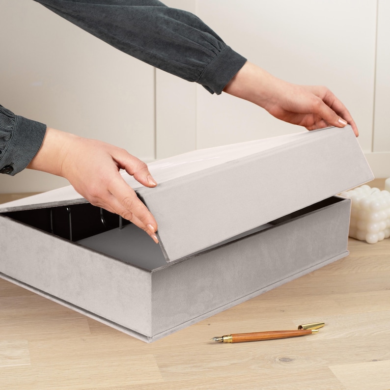 K&ouml;nnte beinhalten: Eine hellgraue rechteckige Aufbewahrungsbox mit passendem Deckel wird ge&ouml;ffnet. Die Box besteht aus einem weichen, samtigen Material. Ein Holzstift mit goldenen Akzenten liegt auf der Holzoberfl&auml;che.