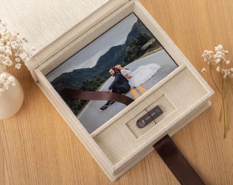 May include: A beige linen-covered box with a hinged lid containing a rectangular photograph of a couple on a road and a silver Philips USB flash drive. A dark brown ribbon is attached to the box for opening and securing the contents inside.