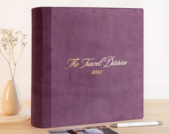 May include: A plush, deep purple travel diary with the gold text "The Travel Diaries 2025". A white pen and a photograph are next to the diary. A small vase with dried flowers is on the left.